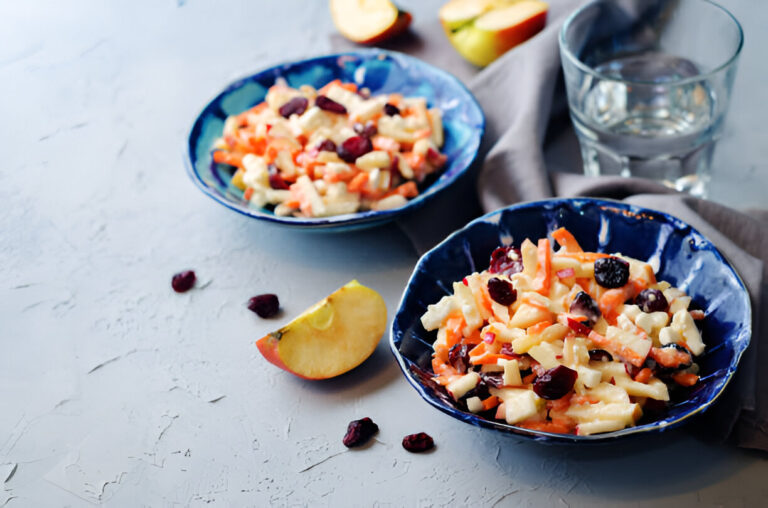 Two bowls of colorful fruit salad with apples, carrots, and cranberries, accompanied by a slice of apple and a glass of water.