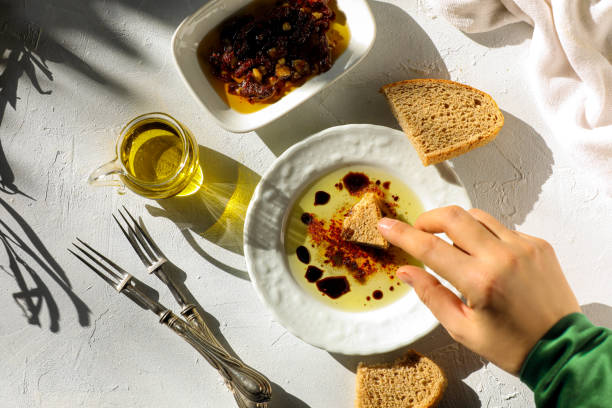 A hand dipping a piece of bread into a bowl of olive oil with spices, accompanied by a small dish of olives and a glass of olive oil.