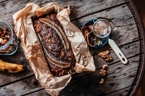 A freshly baked loaf of banana bread in parchment paper, surrounded by nuts and spices on a wooden table.