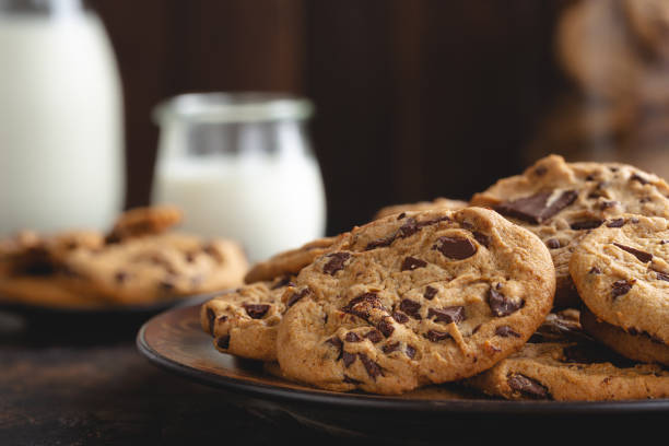 Close-up of freshly baked chocolate chip cookies on a plate with glasses of milk in the background.