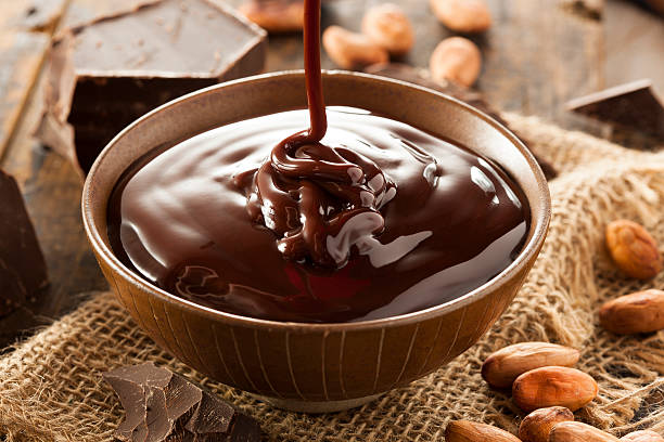 A bowl of rich, melted chocolate with a stream pouring into it, surrounded by cocoa beans and chocolate pieces.