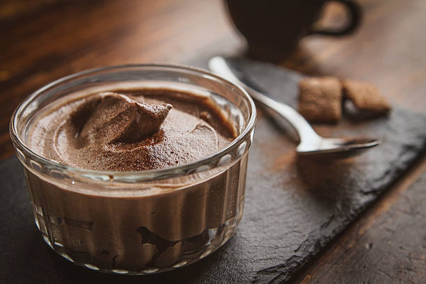 A glass bowl filled with creamy chocolate mousse on a dark slate surface, accompanied by a spoon and some sugar cubes.