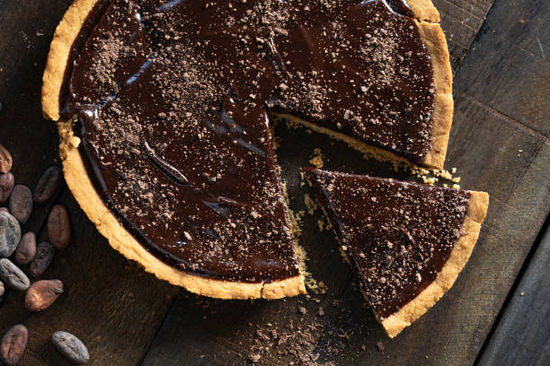 A decadent chocolate tart with a slice cut out, resting on a wooden table surrounded by cocoa beans.