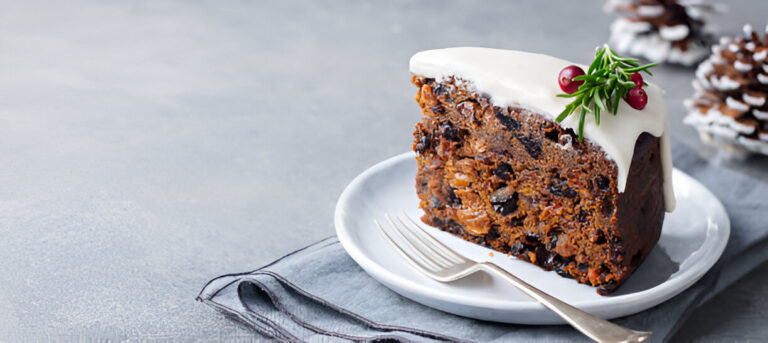 Slice of rich fruitcake topped with white icing and decorative berries on a white plate