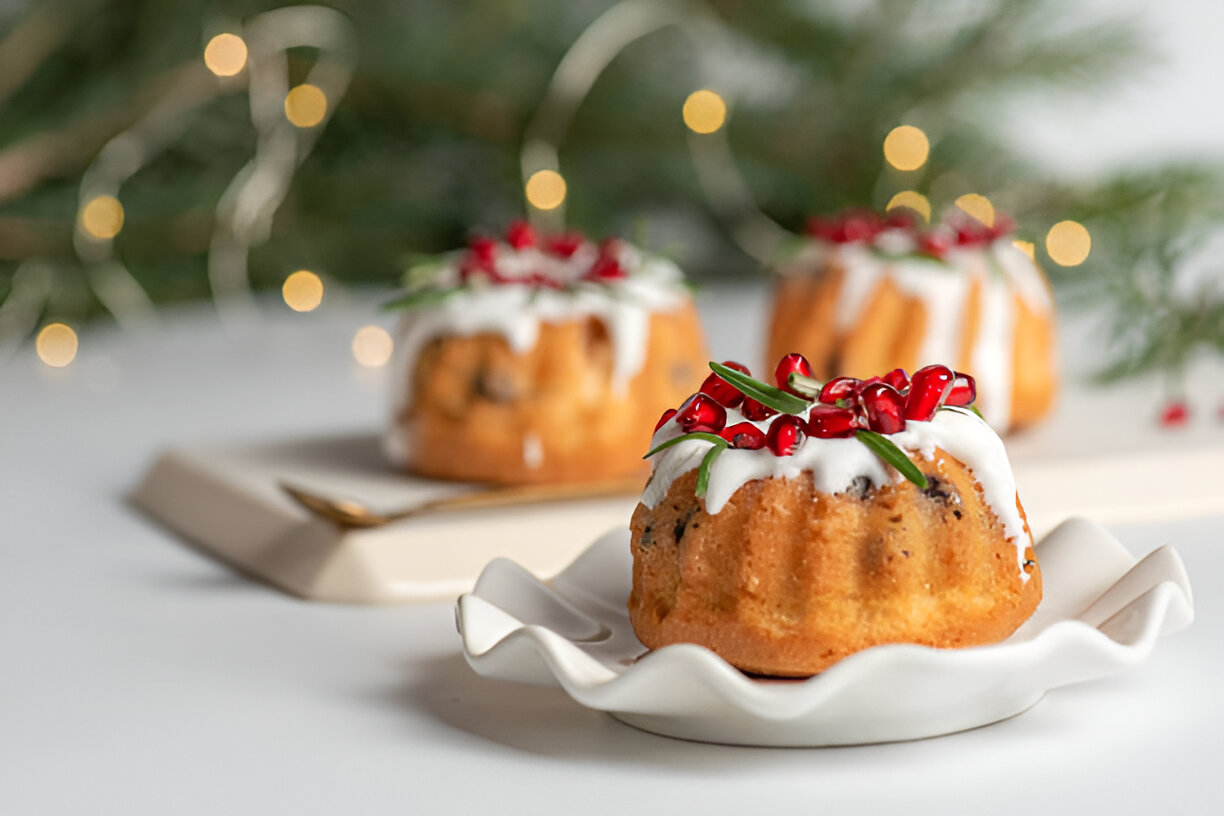 Three mini bundt cakes decorated with icing, pomegranate seeds, and greenery, presented on a white plate with a festive background.