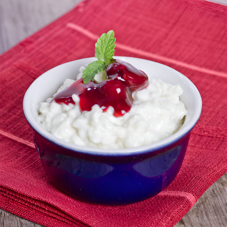 A bowl of creamy cottage cheese topped with red berry jam and a mint leaf on a red cloth background.