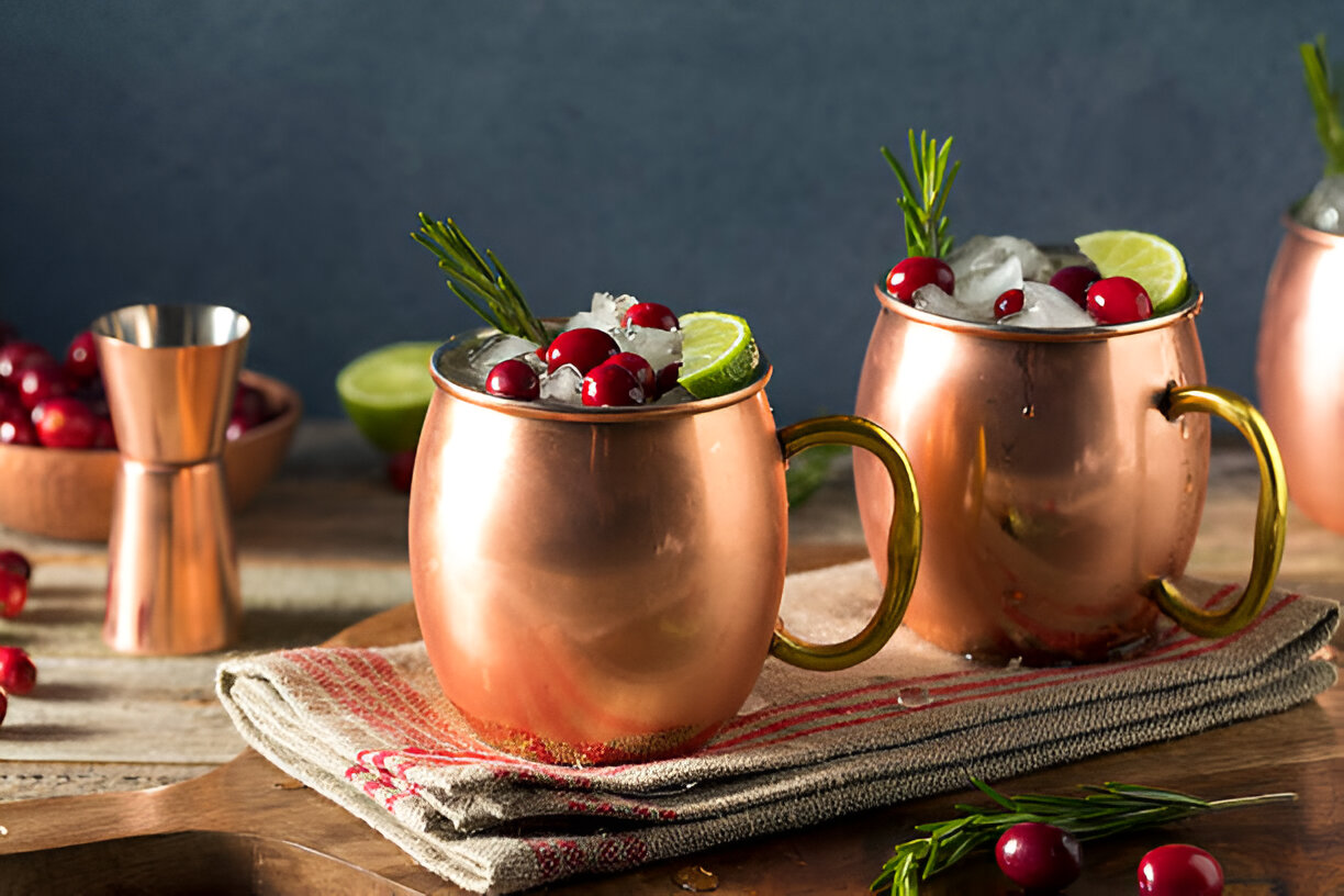 Copper mugs filled with cocktails, garnished with cranberries and lime slices, on a rustic wooden table.