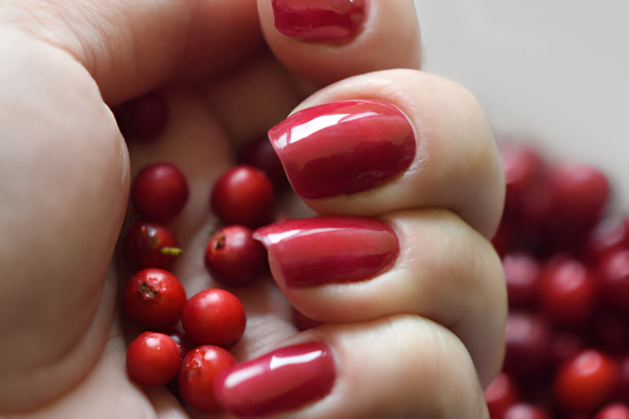 A close-up of a hand holding small red berries with well-manicured nails painted in a glossy red shade.