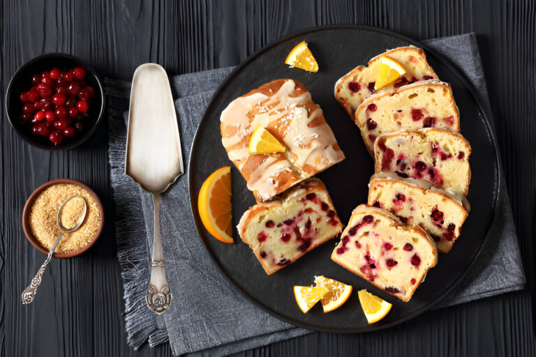 A platter of sliced cranberry bread with orange slices, sugar, and a serving spoon on a dark wooden table.