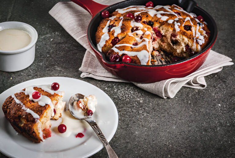 A delicious baked dessert in a red skillet, topped with white icing and fresh cranberries, served on a white plate with a spoon.