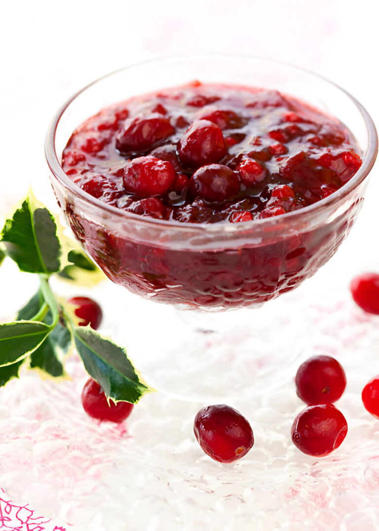 A glass bowl filled with cranberry sauce surrounded by fresh cranberries and green leaves.