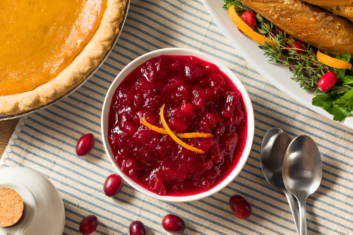 A bowl of cranberry sauce garnished with orange peel next to a pumpkin pie and roasted turkey on a striped tablecloth.