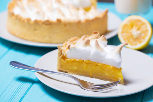 Slice of lemon meringue pie on a white plate with a fork, whole pie in the background, lemon half on the side.