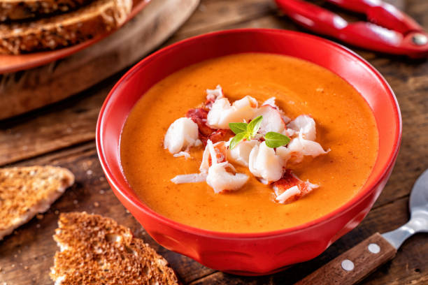 A bowl of creamy lobster bisque topped with fresh lobster meat and herbs, served with toasted bread on a wooden table.