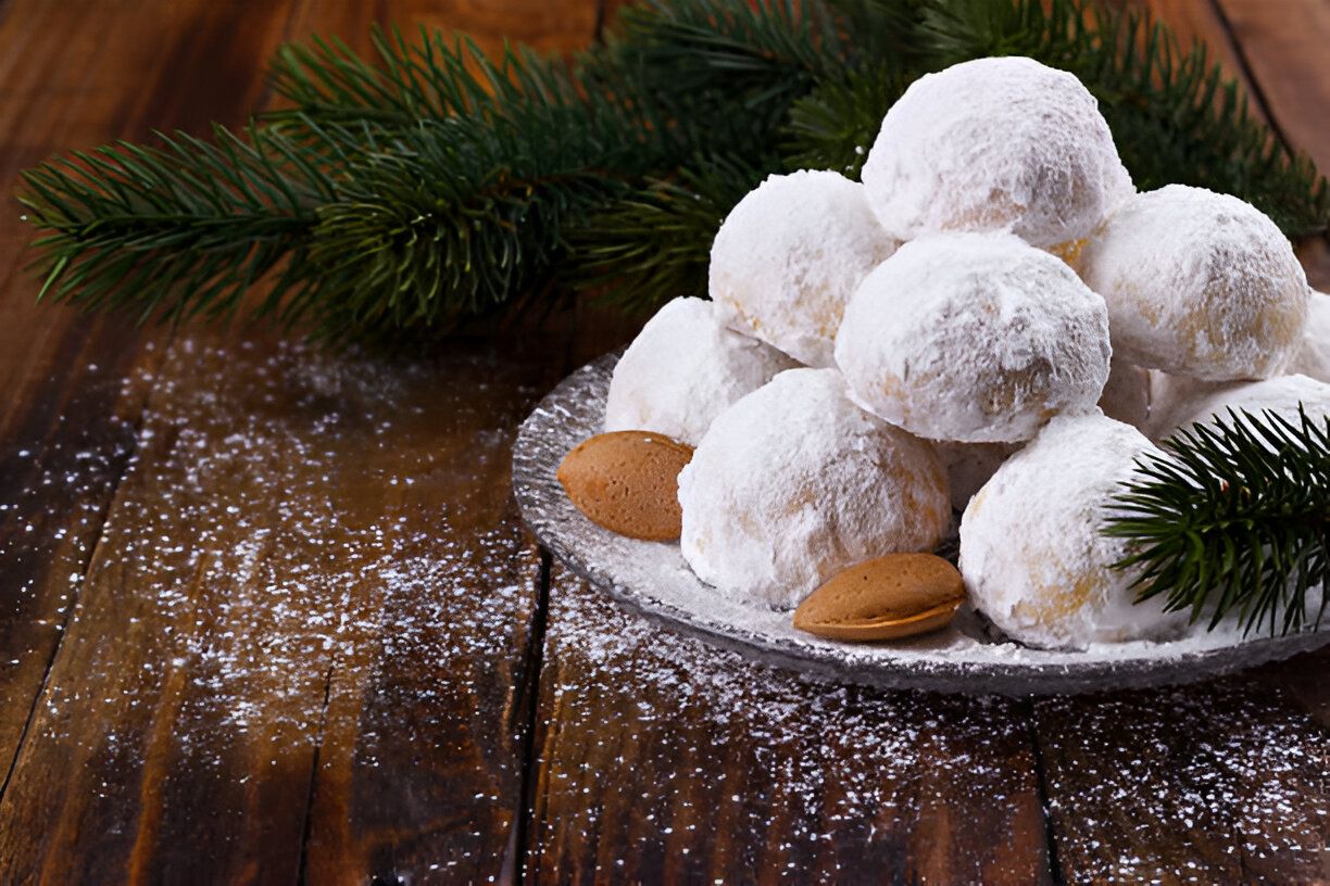 A plate of snowball cookies dusted with powdered sugar, accompanied by gingerbread cookies and pine branches.