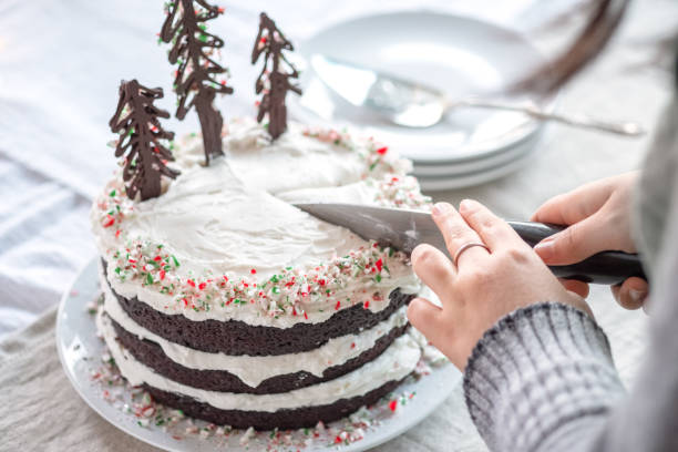 A person slices a decorated three-layer chocolate cake with white frosting and Christmas decorations.