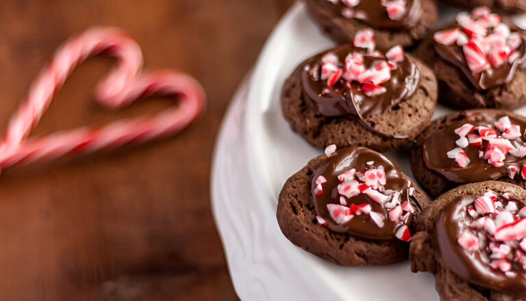 Chocolate cookies topped with peppermint pieces on a white plate, with a blurred candy cane heart in the background.