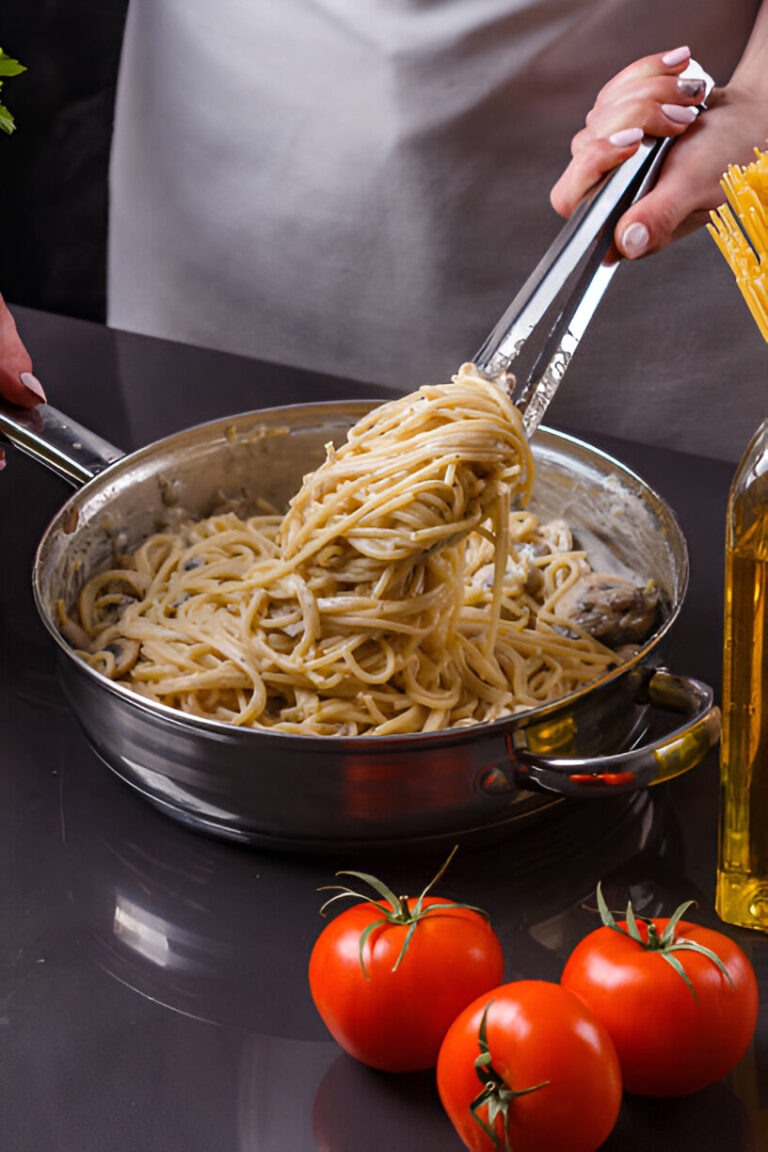 A person using tongs to lift creamy spaghetti from a stainless steel pan, with fresh tomatoes and olive oil in the foreground.