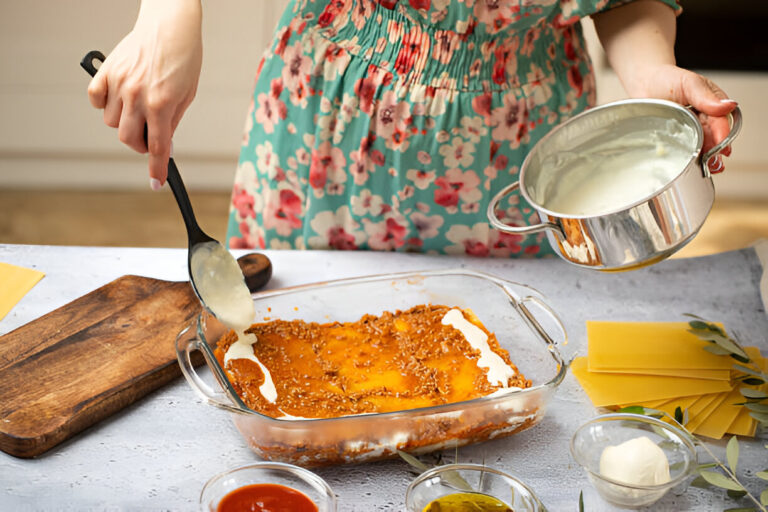 A person preparing a layered dish in a kitchen, pouring creamy sauce into a glass dish that contains pasta and sauce.