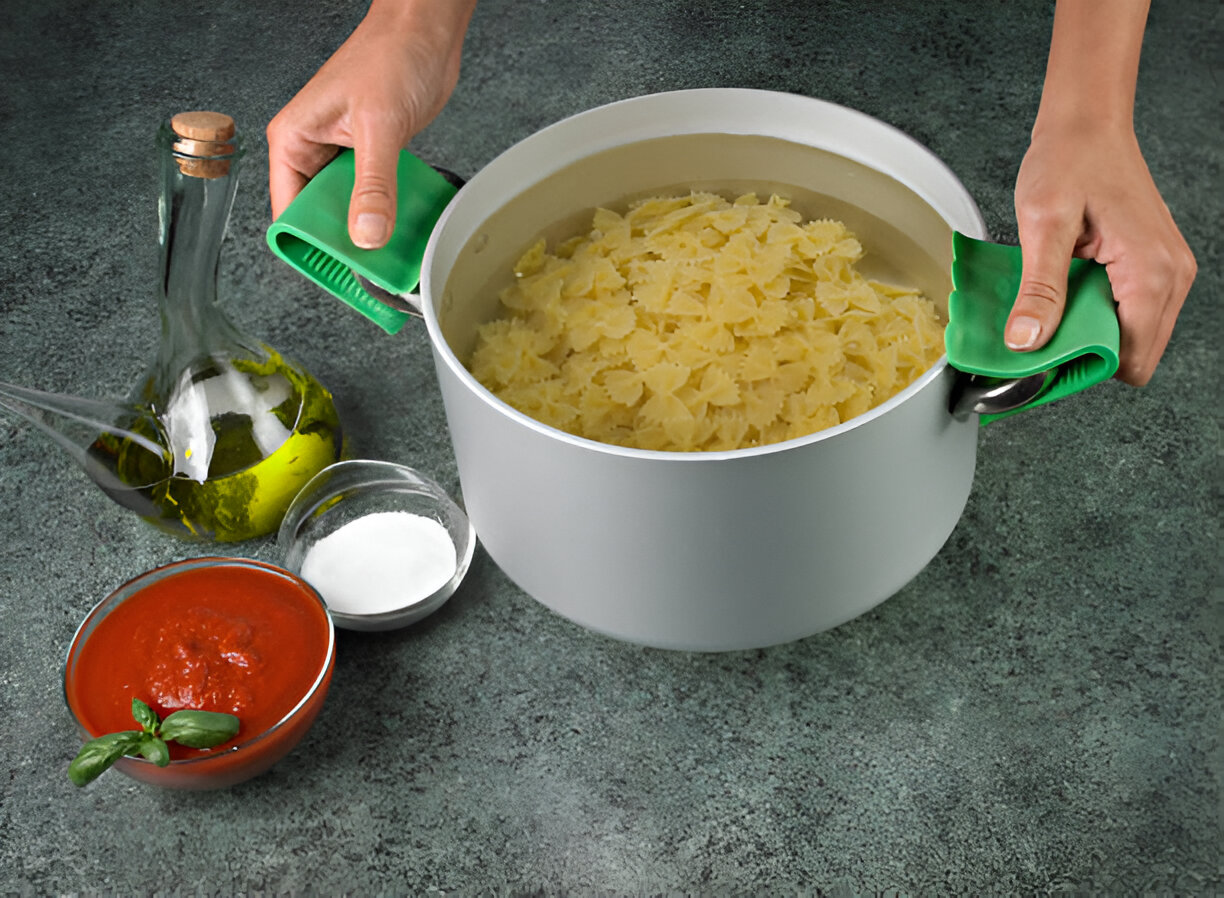 A pot of cooked bowtie pasta being held with green silicone handles, alongside olive oil, tomato sauce, and salt on a textured surface.