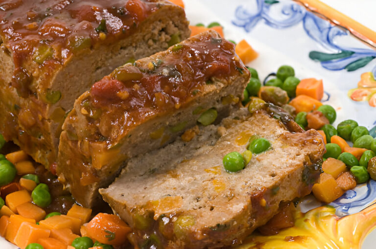 A slice of meatloaf topped with a tangy sauce, surrounded by peas and diced carrots on a decorative plate.