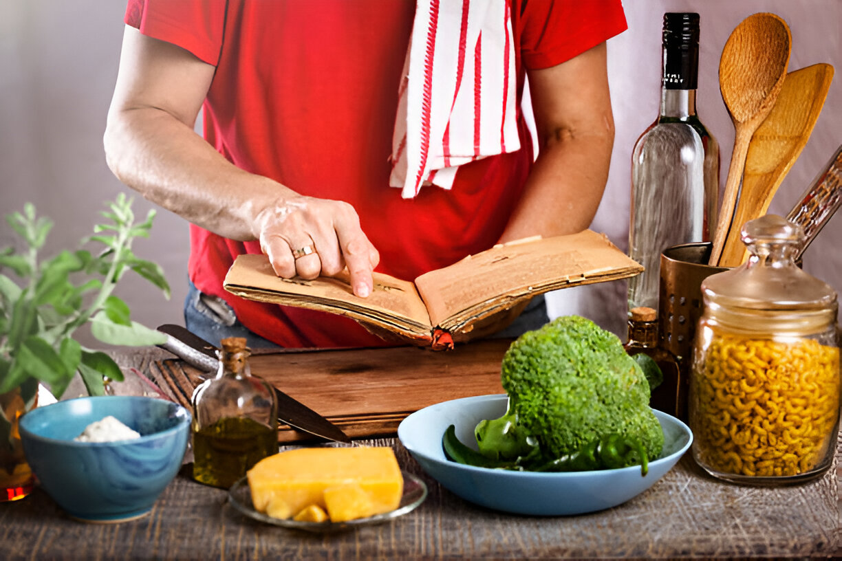 A person in a red shirt pointing at an open recipe book in a rustic kitchen setting, surrounded by fresh ingredients.