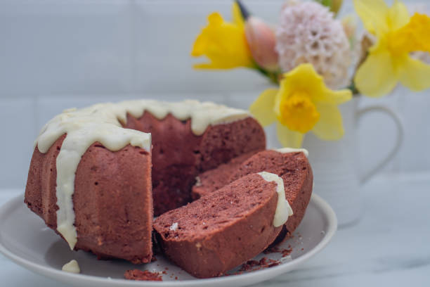 A slice of chocolate bundt cake with white icing on a plate, alongside a vase of colorful flowers.