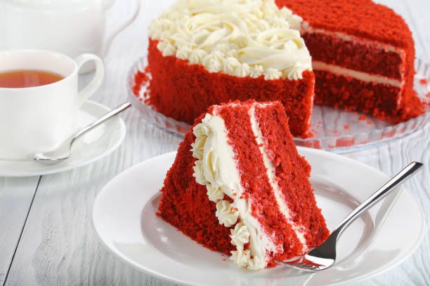 A slice of red velvet cake with cream cheese frosting on a white plate, next to a cup of tea.