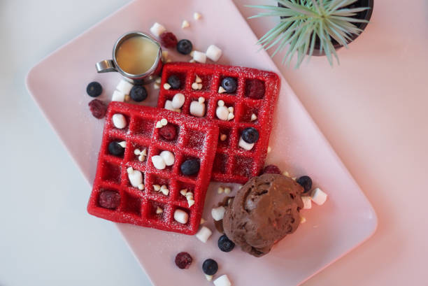 Two red waffles garnished with blueberries, raspberries, mini marshmallows, and a scoop of chocolate ice cream served with a small jug of syrup.