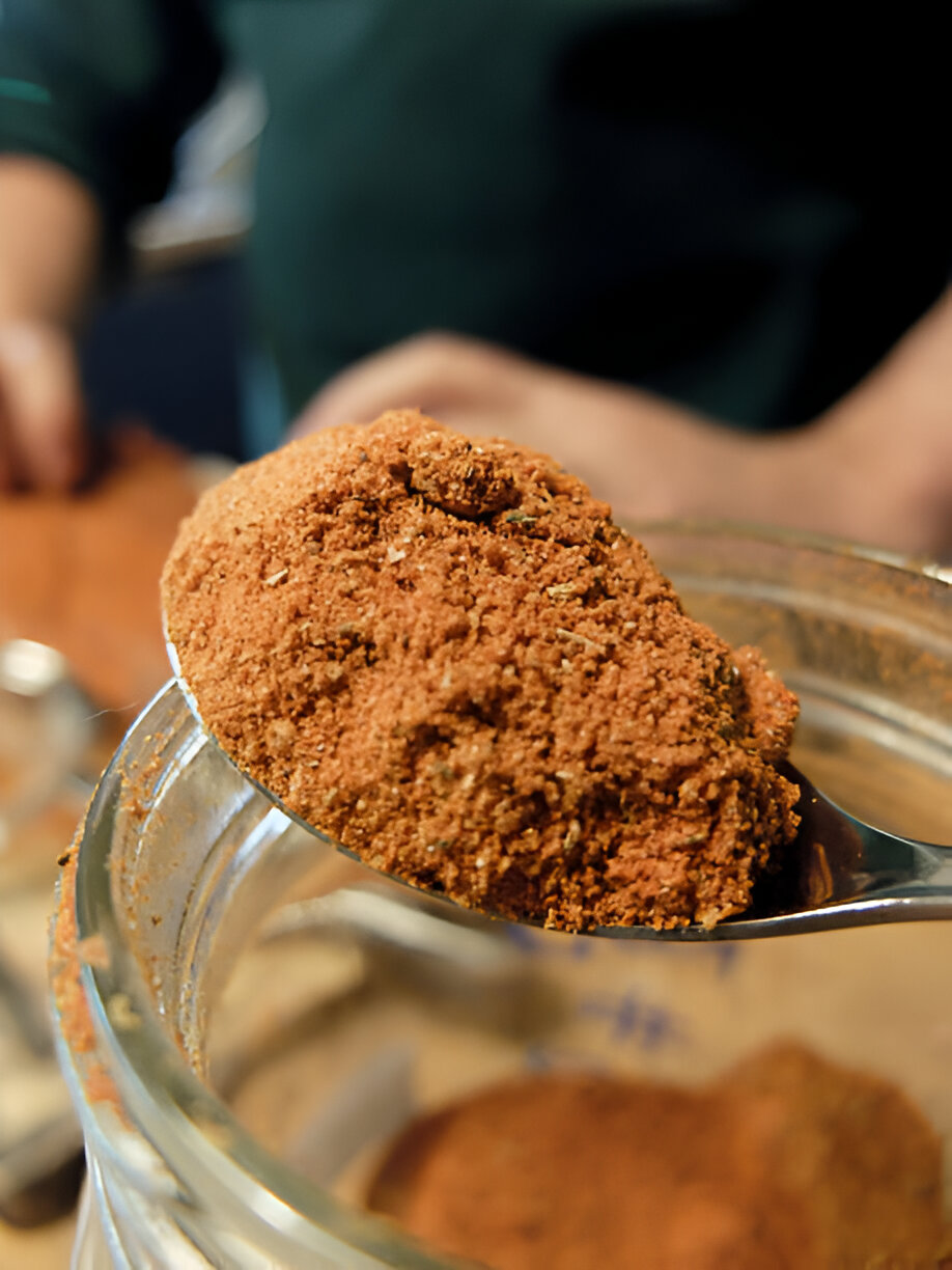 A spoonful of reddish-brown powder being lifted from a glass jar.