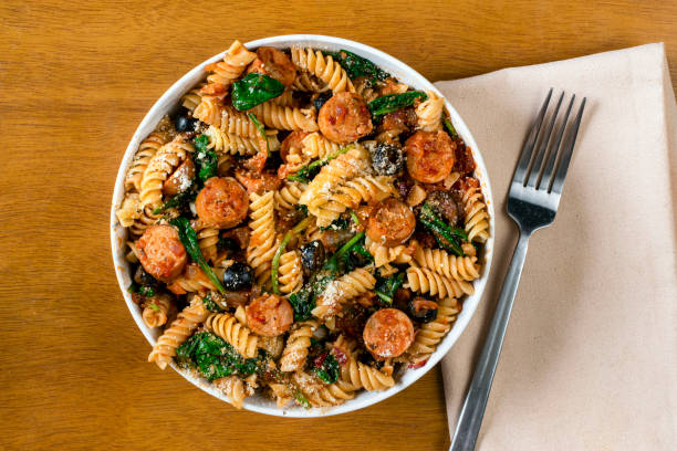 A bowl of fusilli pasta with sausage, spinach, and olives, served on a wooden table with a fork and a napkin.