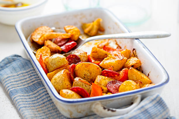 A baked dish of roasted potatoes and sausage served in a white dish with a silver spoon.