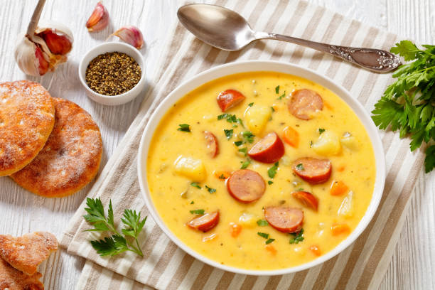 A bowl of creamy sausage soup with potatoes and vegetables, accompanied by garlic, herbs, and round biscuits on a striped napkin.