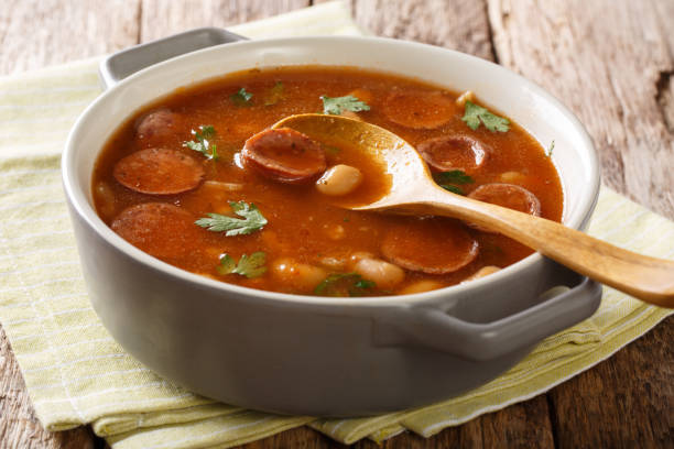 A bowl of hearty stew with sliced sausage and leafy greens on a wooden table.