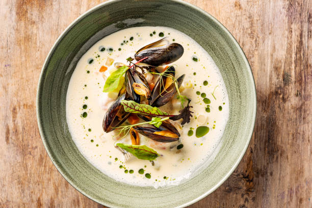 A bowl of creamy mussel soup garnished with fresh herbs and vibrant green oil drizzles, resting on a rustic wooden table.