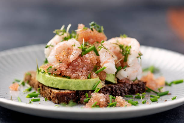 A gourmet dish featuring avocado and shrimp on dark bread, garnished with fish roe and chopped herbs.