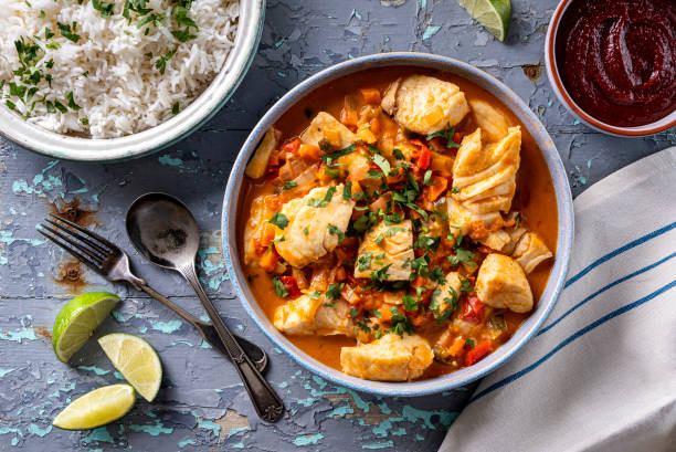 A vibrant bowl of fish stew with peppers and herbs, accompanied by a side of fluffy white rice and lime wedges.