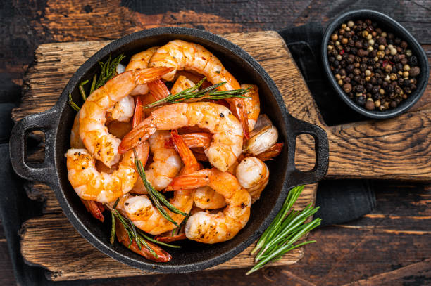 A skillet filled with cooked shrimp garnished with rosemary on a wooden board, accompanied by a bowl of black peppercorns.