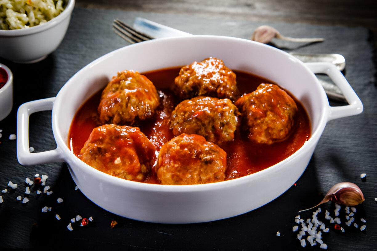 A white dish filled with meatballs in red sauce, garnished with spices and garlic.