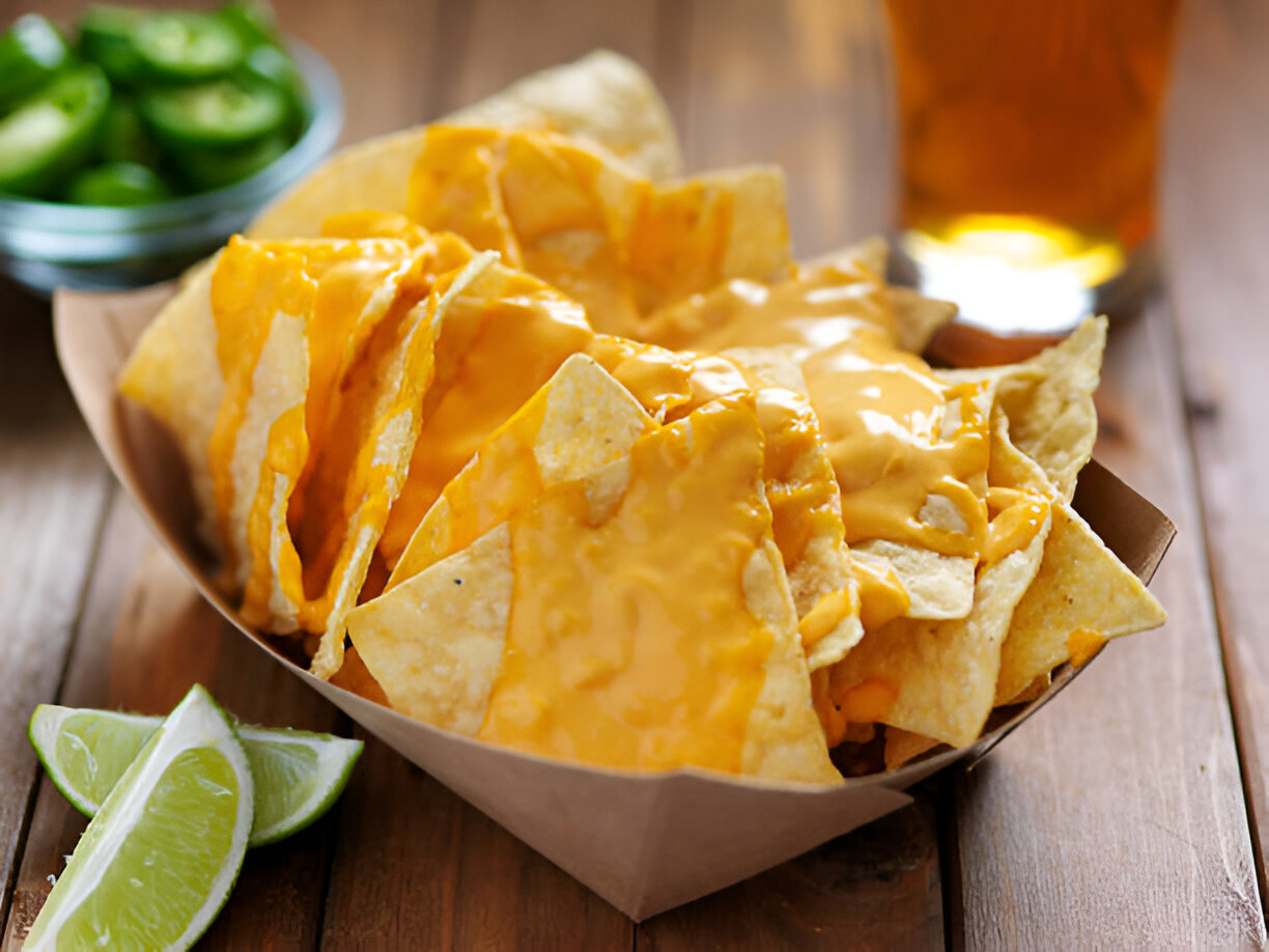 A serving of cheesy nachos with lime wedges and jalapeños in the background.