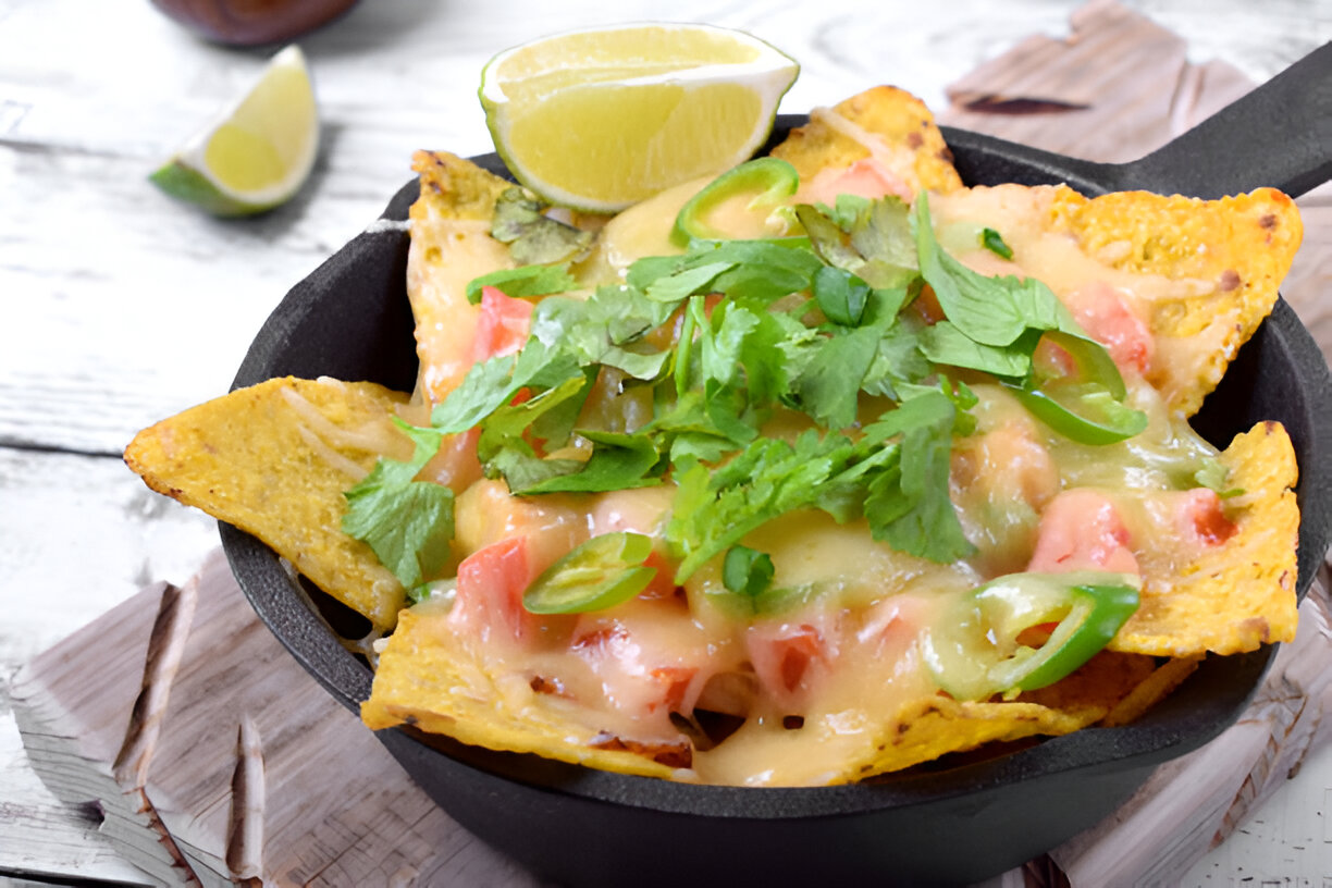 A delicious serving of nachos topped with melted cheese, diced tomatoes, green onions, and cilantro, served in a cast iron skillet with lime wedges on the side.