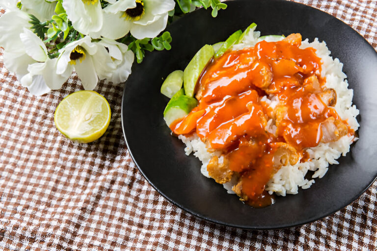 A plate of rice topped with fried chicken covered in sauce, accompanied by lime slices and flowers.