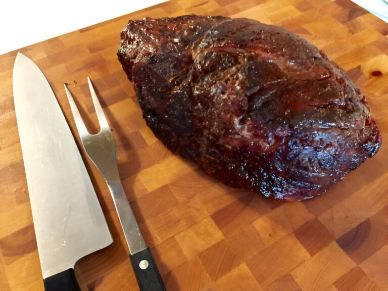 A large piece of cooked meat on a wooden cutting board, accompanied by a knife and a carving fork.