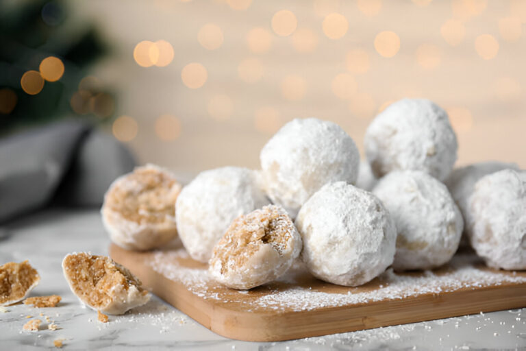 A close-up of powdered sugar-covered dessert balls on a wooden cutting board, with one partially bitten into and crumbs scattered nearby.