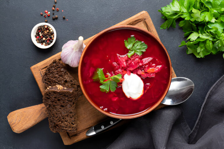 A bowl of vibrant red borscht soup garnished with parsley and sour cream, served with slices of dark bread and garlic on a wooden board.