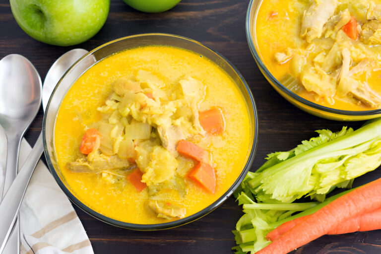 Two bowls of chicken soup with vegetables on a wooden surface, surrounded by fresh greens and carrots.