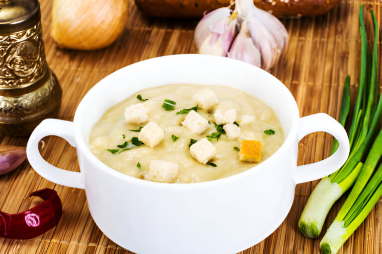 A bowl of creamy soup topped with croutons and herbs, with garlic and onions in the background.