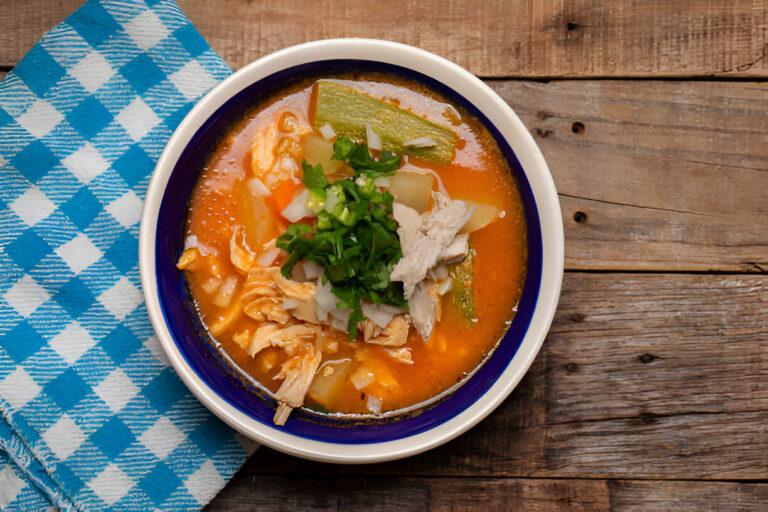 A bowl of soup with chicken, vegetables, and green herbs on a wooden table with a blue checkered towel.