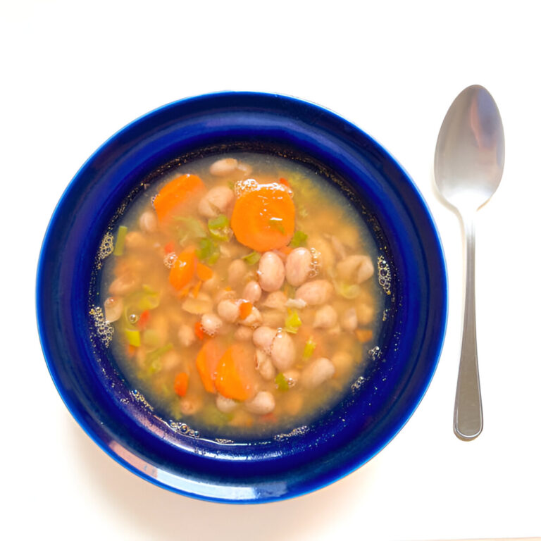 A blue bowl filled with soup containing beans, carrots, and herbs, beside a silver spoon on a white background.