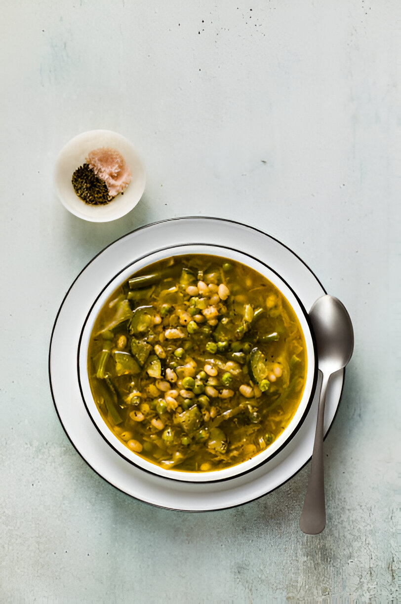 A bowl of green vegetable soup with beans on a pale surface, accompanied by a small dish of spices.